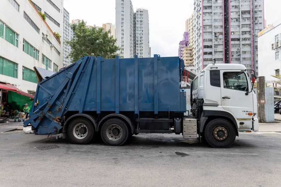 A blue garbage truck parked in a vibrant city street surrounded by tall buildings.